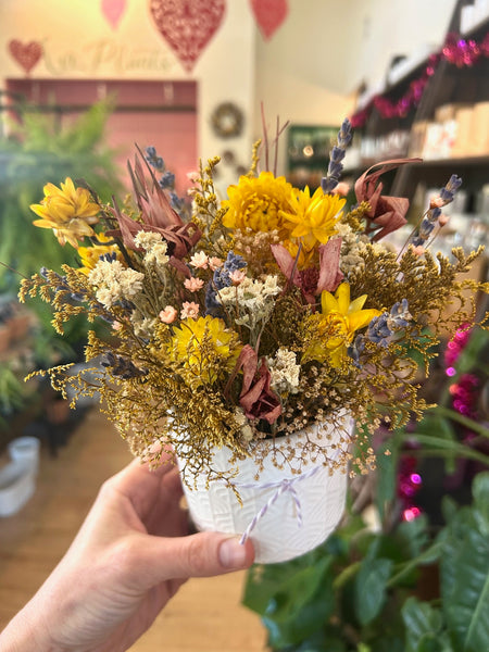 Hand holding a small bouquet of dried flowers in a white pot with a blurred indoor background.